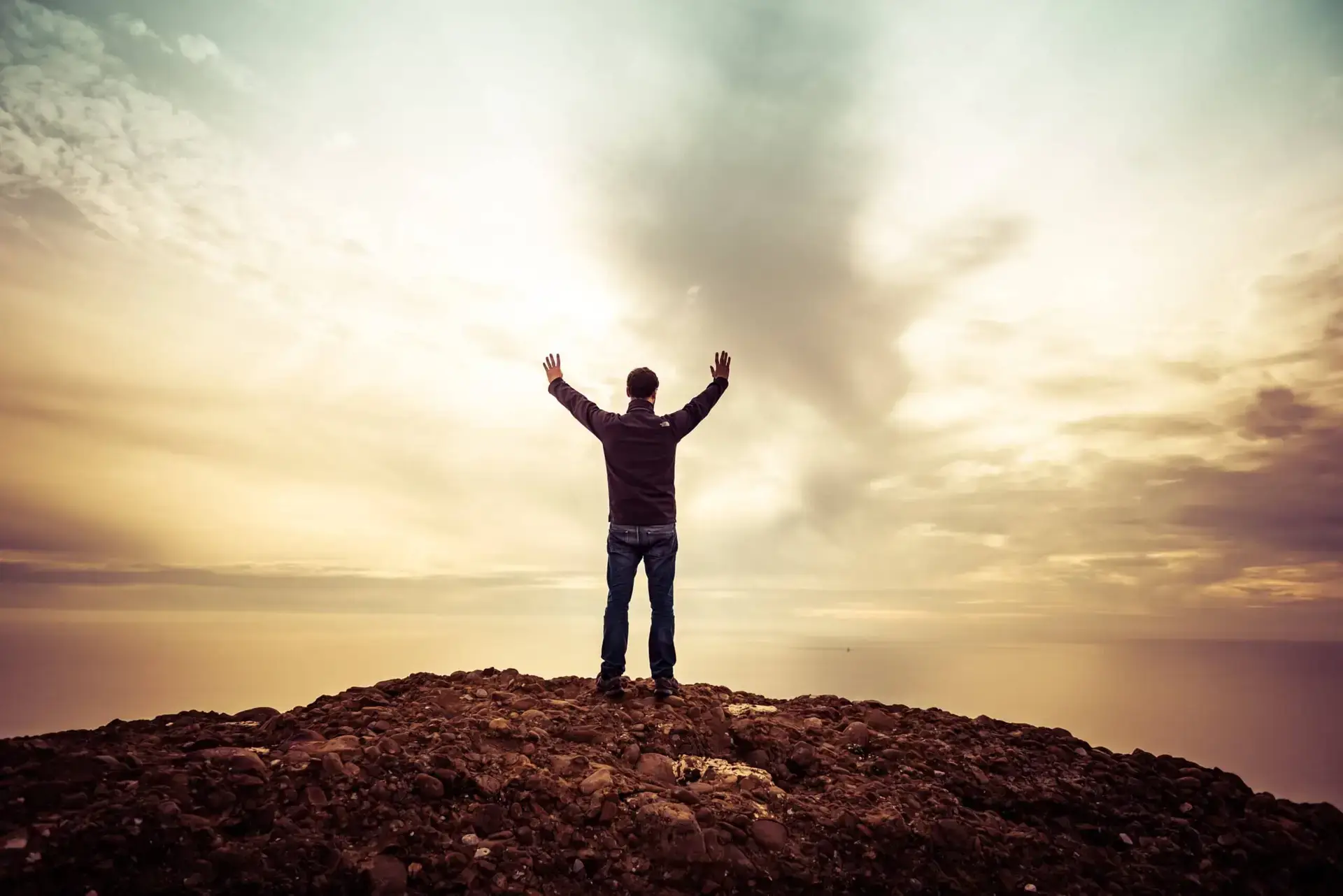 Man standing on rocks overlooking water with arms raised in worship