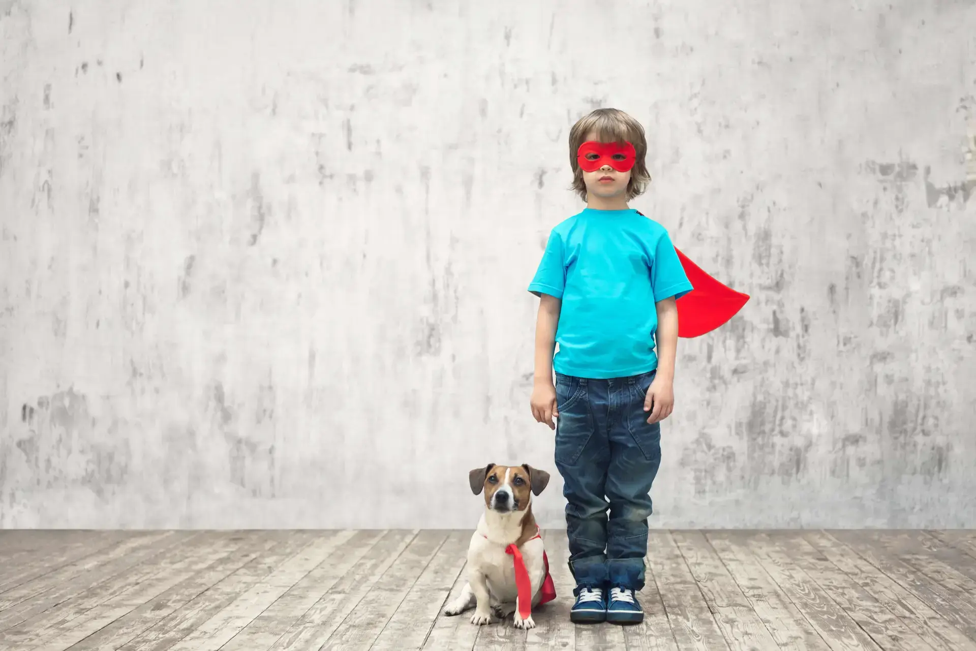 Little boy in super hero cape and mask with dog beside him also wearing a cape