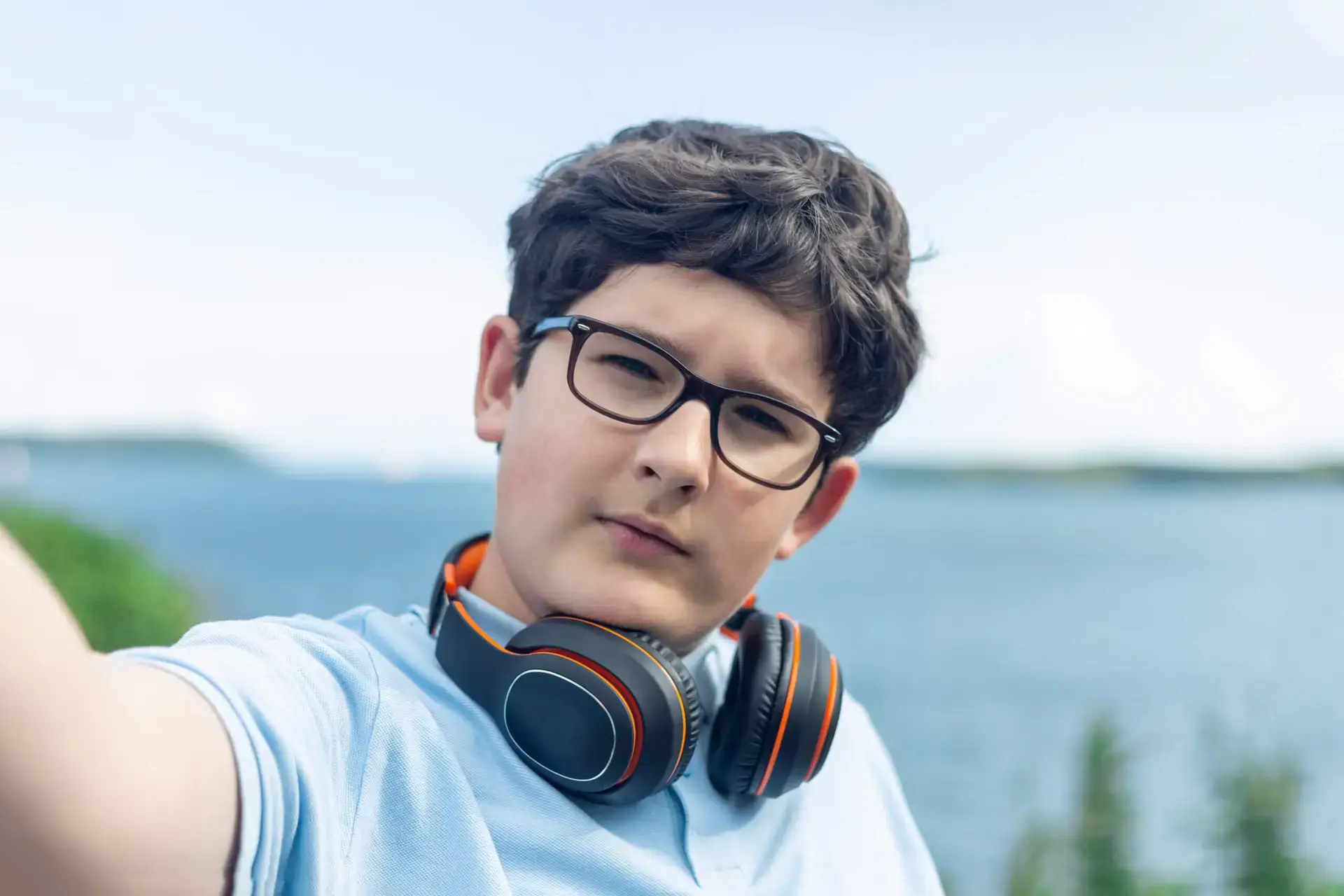 Portrait of eleven year old boy in glasses and wireless headphones, taking a selfie in front of sea