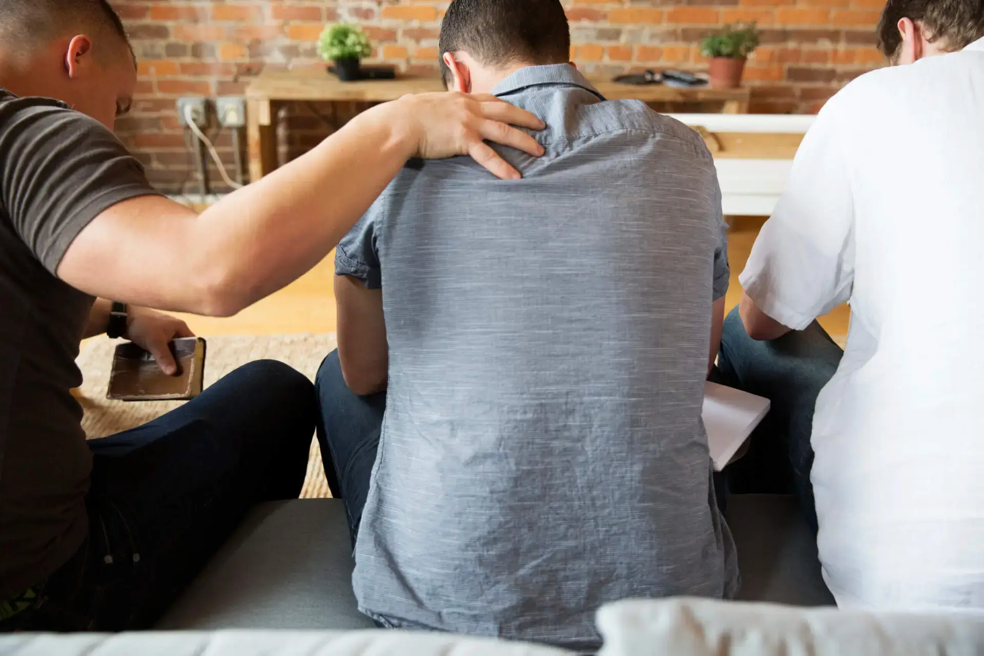 Three men sitting together on a couch praying with man on left placing hand on the back of man in the center