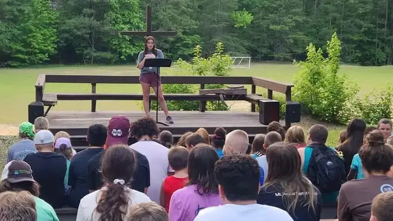 Young woman stands on outdoor stage in front of audience at WorldSong Camp