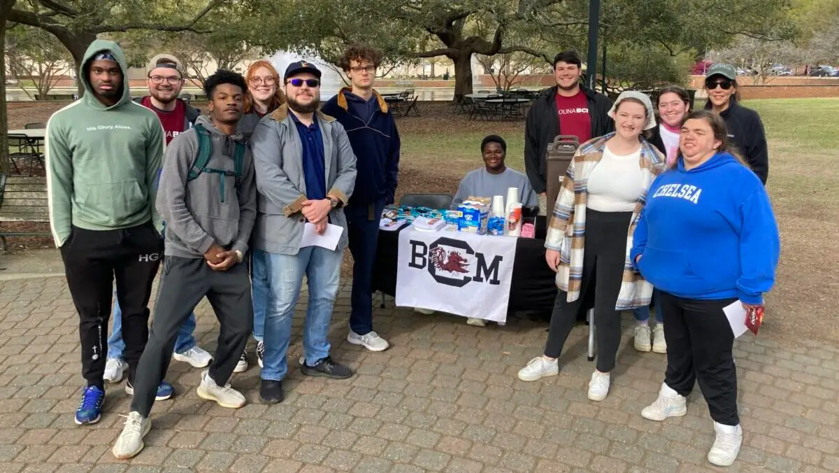 A large group of college students stand on either side of a table with a BCM table cloth on the quad at the University of South Carolina