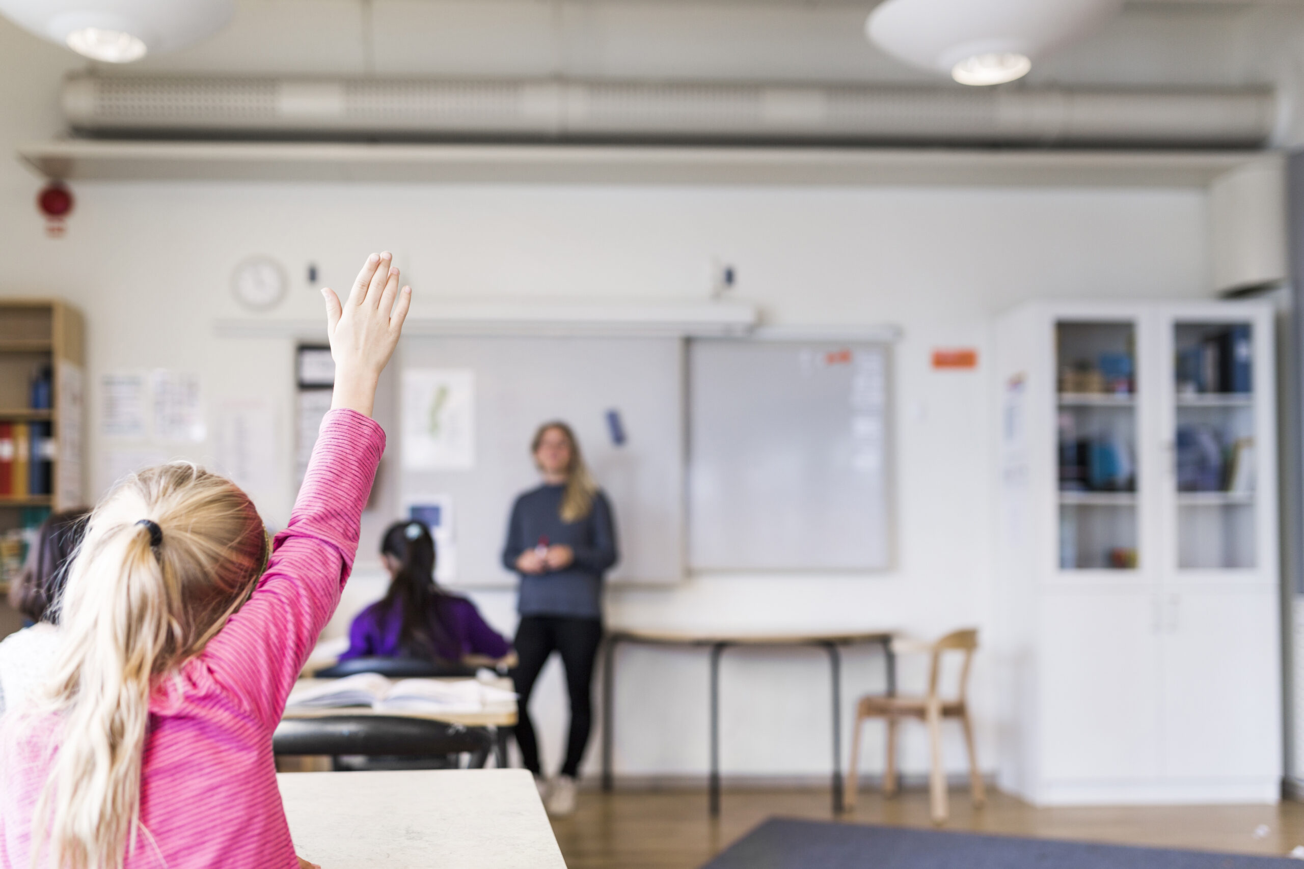 Eager student raising hand in classroom with teacher in the background.