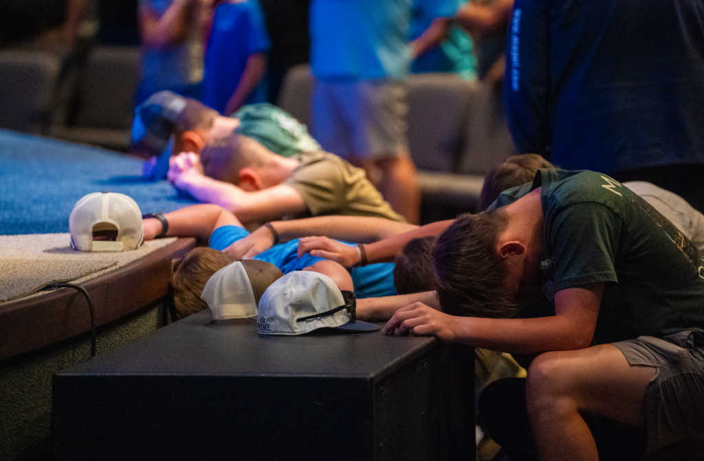 Teenage guys kneel at a church stage altar with hats sitting on the stage and hands on each other's backs in prayer