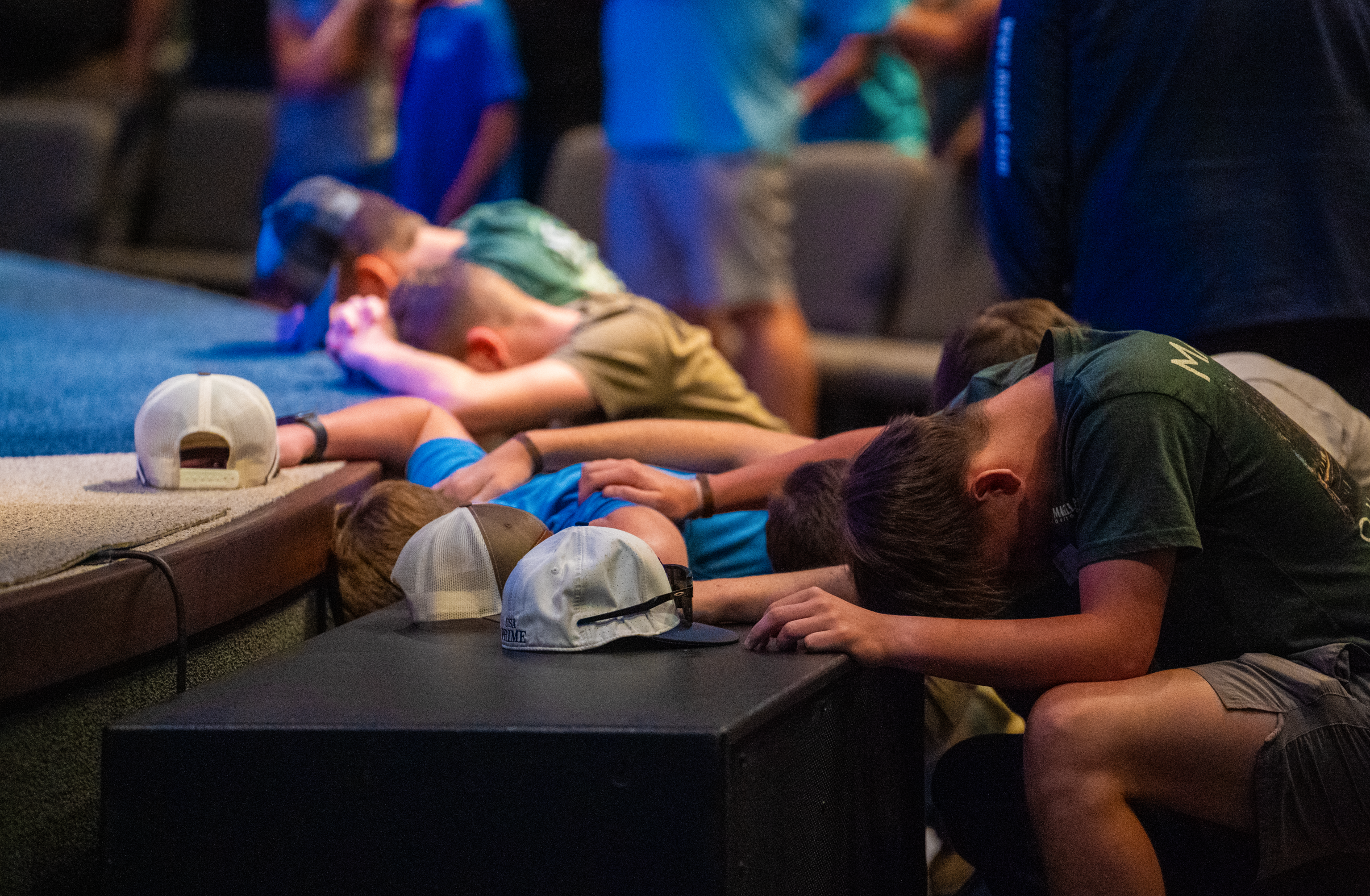 Teenage guys kneel at a church stage altar with hats sitting on the stage and hands on each other's backs in prayer